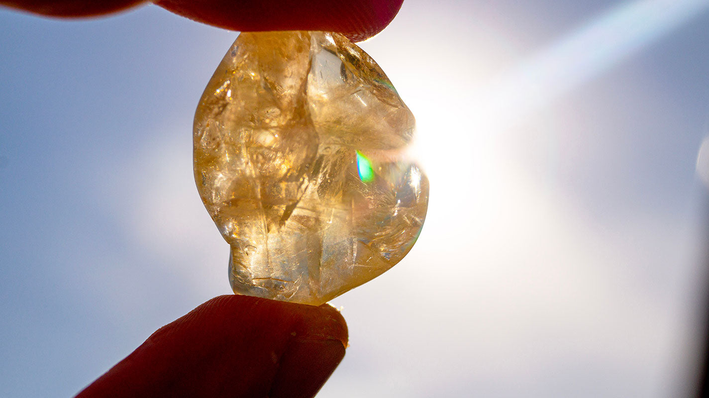 A close-up of a raw citrine gemstone held between two fingers against the sunlight. The golden-yellow crystal glows as the light passes through, revealing its natural inclusions and textures.