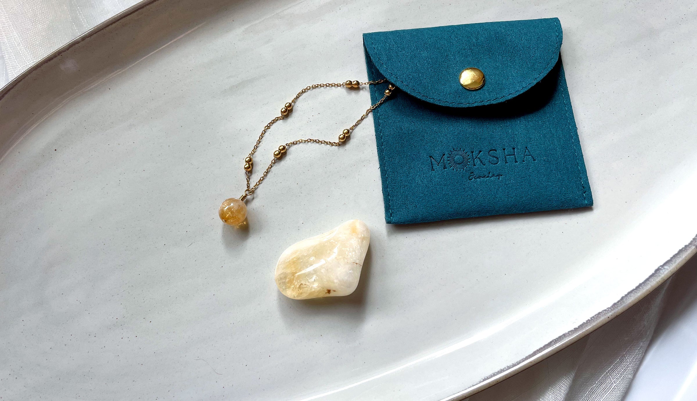 A delicate gold chain with small gold balls and a 10mm Citrine bead pulled out of an emerald pouch with a Moksha Jewelry Logo embossing next to a Citrine gemstone on a gray ceramic plate.