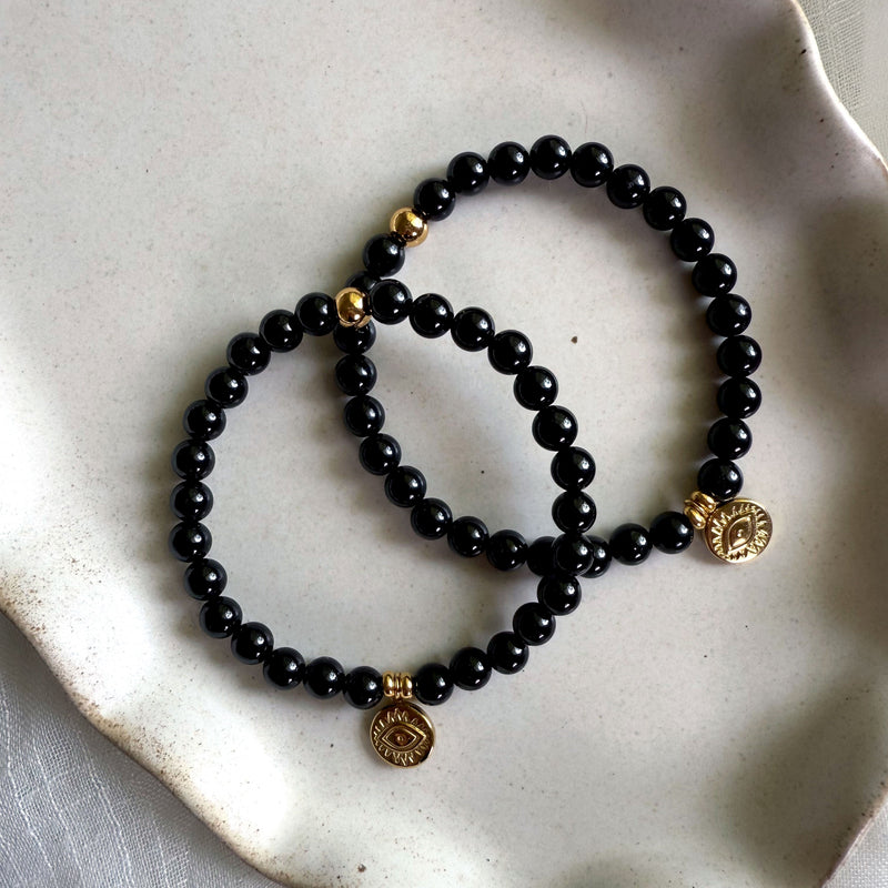 Pair of black tourmaline gemstone bracelets with gold evil eye charms displayed on neutral-toned ceramic dish. Handmade matching protection jewelry set.
