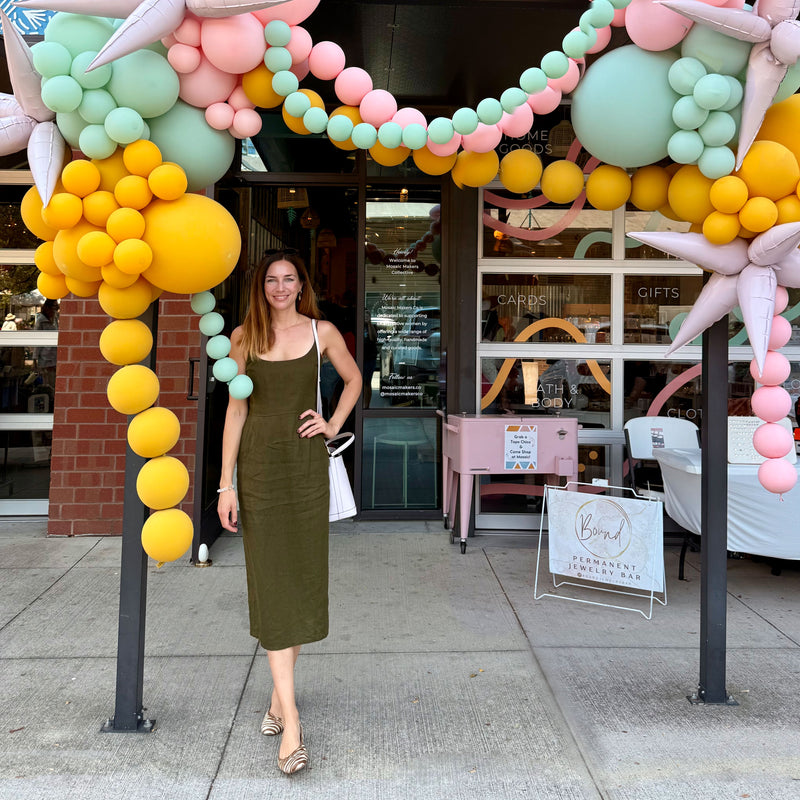 Vanessa Simion-Mueller, founder of Moksha Jewelry, standing in front of the Mosaic Makers Collective Store at Dallas Farmers Market. The storefront is decorated with colorful pastel balloon garlands celebrating the grand opening in September 2025.