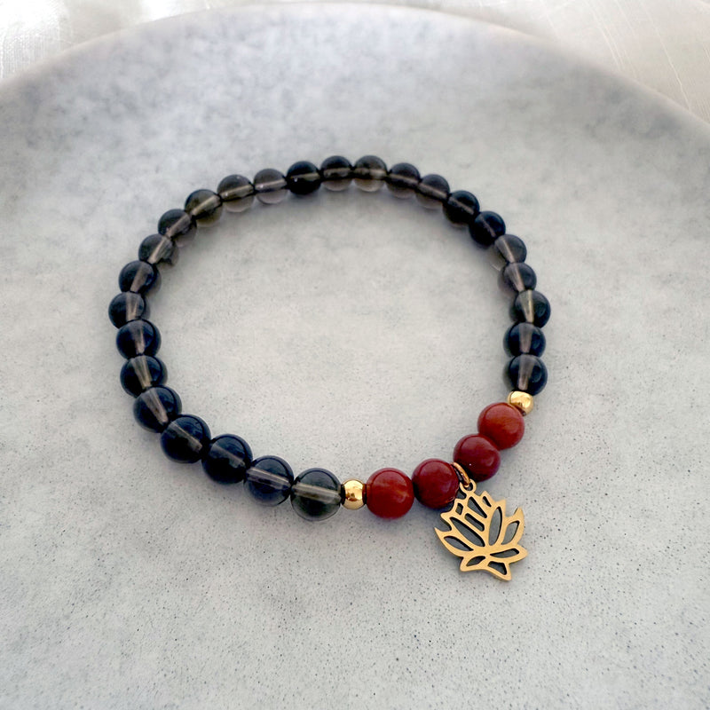 Chakra bracelet made with smoky quartz and red jasper beads, featuring a gold-plated lotus charm and gold accents, handmade by Moksha Jewelry and displayed on a neutral stone background.