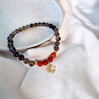 Handmade root chakra bracelet featuring smoky quartz and red jasper beads with gold accents and a gold-plated lotus charm, crafted by Moksha Jewelry and styled on a soft neutral fabric with a ceramic plate for a serene, natural look.