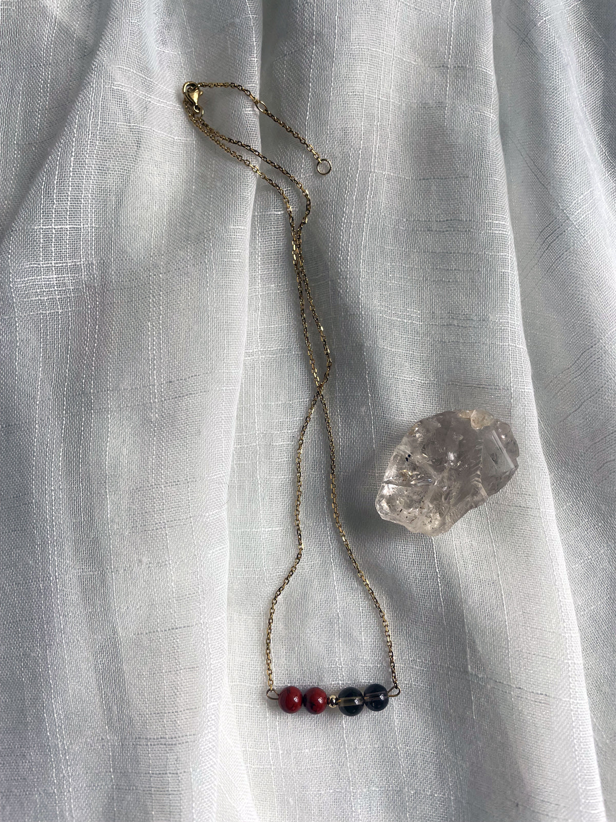 A fine golden necklace with 2 red jasper beads, 2 smoky quartz beads and one golden bead in the middle on a light fabric, next to a raw smoky quartz gemstone.