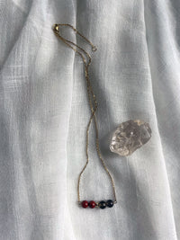 A fine golden necklace with 2 red jasper beads, 2 smoky quartz beads and one golden bead in the middle on a light fabric, next to a raw smoky quartz gemstone.