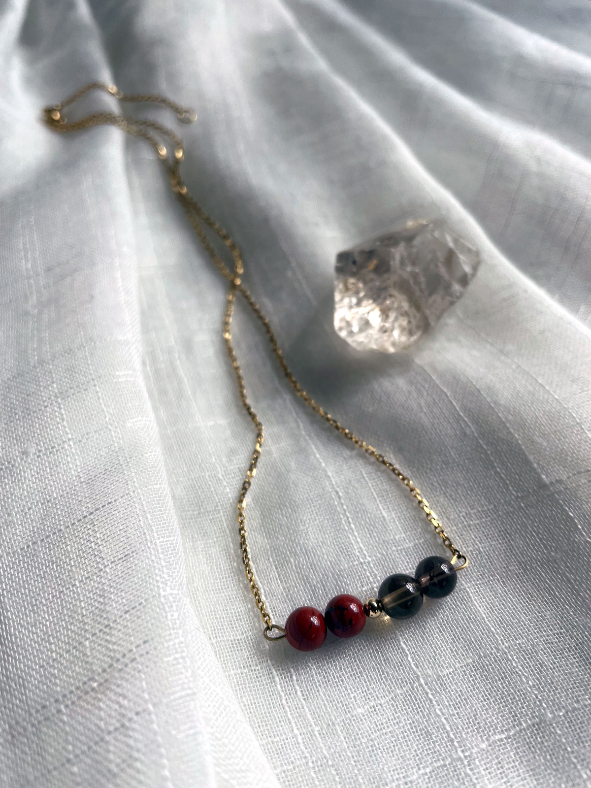 A close-up of a fine golden necklace with 2 red jasper beads, 2 smoky quartz beads and one golden bead in the middle on a light fabric, next to a raw smoky quartz gemstone.