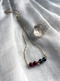 A close-up of a fine golden necklace with 2 red jasper beads, 2 smoky quartz beads and one golden bead in the middle on a light fabric, next to a raw smoky quartz gemstone.
