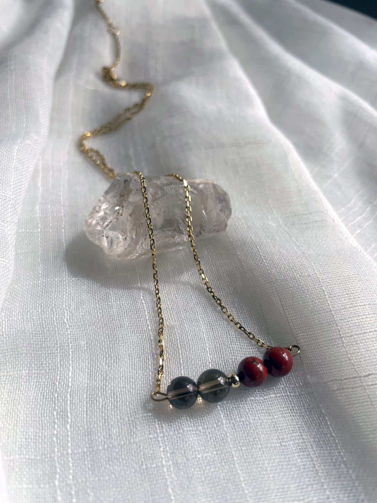 A fine golden necklace with 2 red jasper beads, 2 smoky quartz beads and one golden bead in the middle on a light fabric, lying over a raw smoky quartz gemstone.
