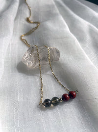 A fine golden necklace with 2 red jasper beads, 2 smoky quartz beads and one golden bead in the middle on a light fabric, lying over a raw smoky quartz gemstone.