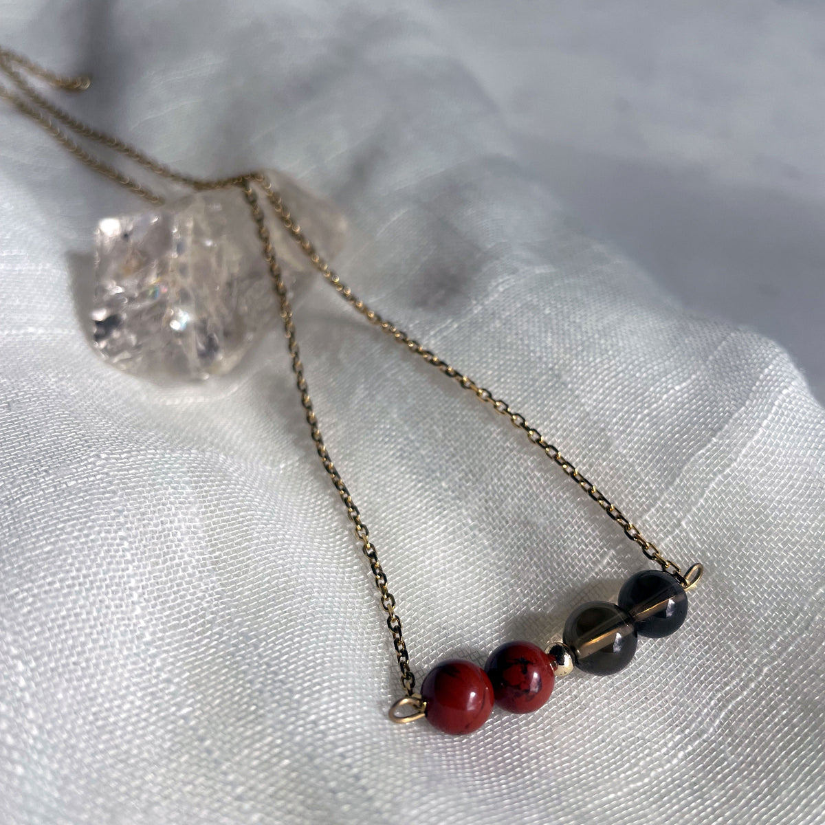 A close-up of a fine golden necklace with 2 red jasper beads, 2 smoky quartz beads and one golden bead on a light fabric, lying over a raw smoky quartz gemstone.