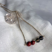 A close-up of a fine golden necklace with 2 red jasper beads, 2 smoky quartz beads and one golden bead on a light fabric, lying over a raw smoky quartz gemstone.