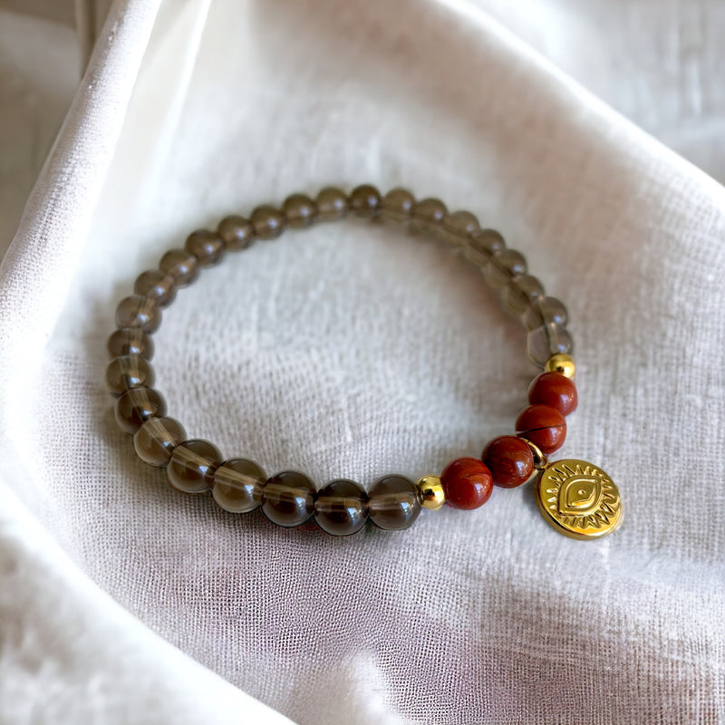 A bracelet made of smoky quartz beads, 4 red jasper beads and 2 small golden beads with a golden evil eye charm in the middle on a light brown fabric.