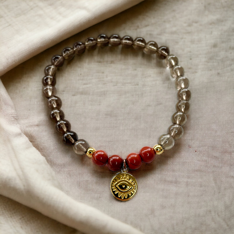 A bracelet made of smoky quartz beads, 4 red jasper beads and 2 small golden beads with a golden evil eye charm in the middle on a light brown fabric.