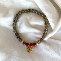 A bracelet made of smoky quartz beads, 4 red jasper beads and 2 small golden beads with a golden evil eye charm in the middle on a light brown fabric.