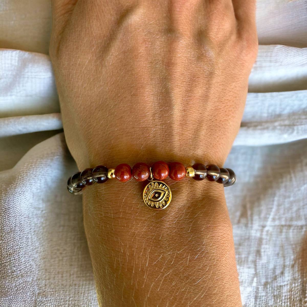 A bracelet made of smoky quartz beads, 4 red jasper beads and 2 small golden beads with a golden evil eye charm in the middle worn on a wrist on a light brown fabric.