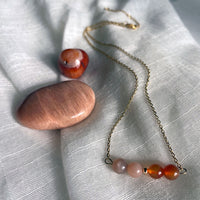 A close-up of a fine golden necklace with 2 carnelian beads, 2 moonstone beads and one golden bead in the middle on a light fabric. Next to the necklace is a carnelian and a moonstone gemstone.