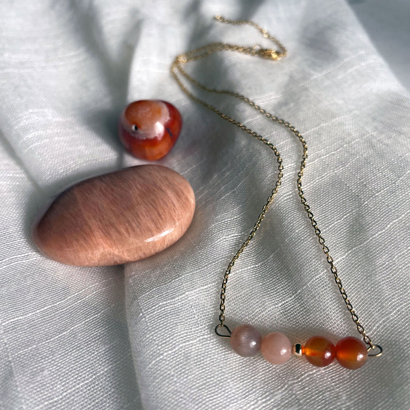 A close-up of a fine golden necklace with 2 carnelian beads, 2 moonstone beads and one golden bead in the middle on a light fabric. Next to the necklace is a carnelian and a moonstone gemstone.