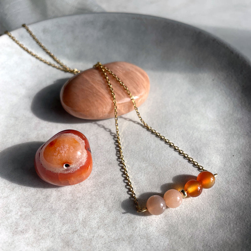A close-up of a fine golden necklace with 2 carnelian beads, 2 moonstone beads and one golden bead in the middle on a ceramic plate. Next to the necklace is a carnelian and a moonstone gemstone.
