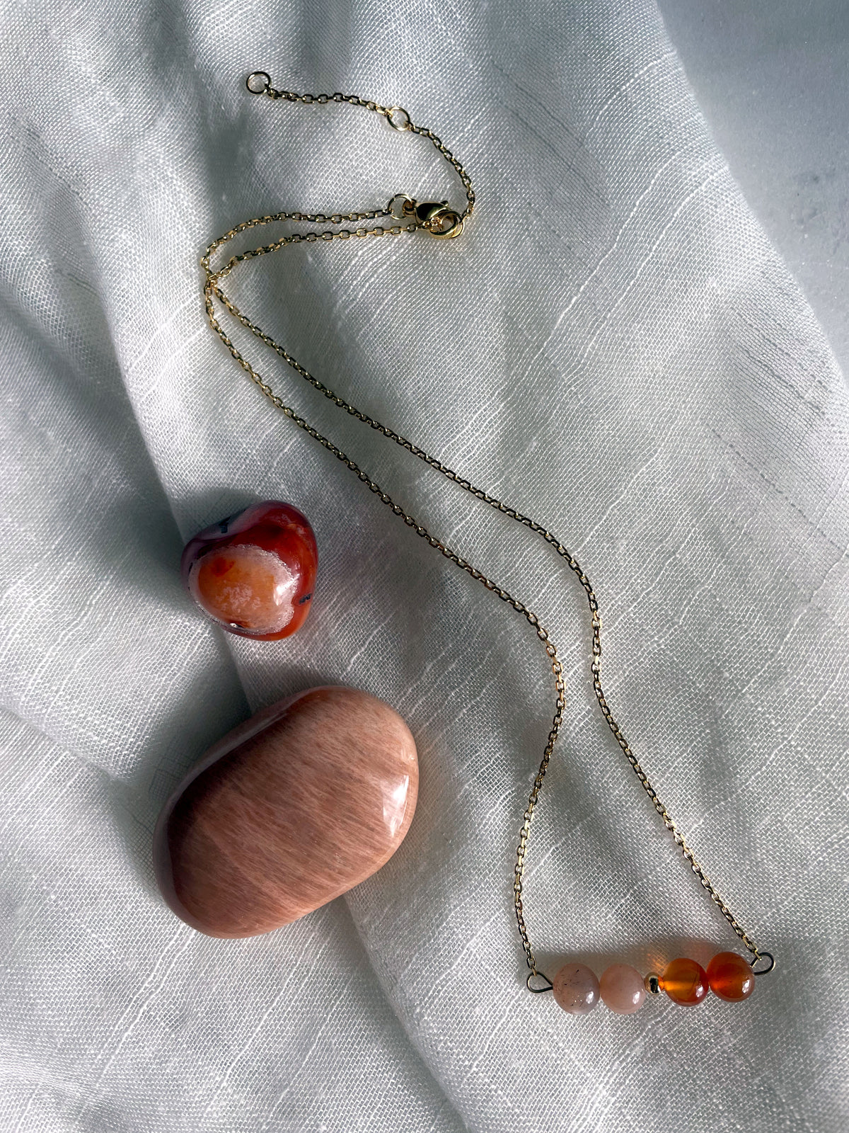 A fine golden necklace with 2 carnelian beads, 2 moonstone beads and one golden bead in the middle on a light fabric. Next to the necklace is a carnelian and a moonstone gemstone.