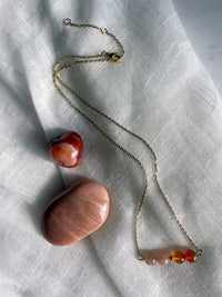 A fine golden necklace with 2 carnelian beads, 2 moonstone beads and one golden bead in the middle on a light fabric. Next to the necklace is a carnelian and a moonstone gemstone.