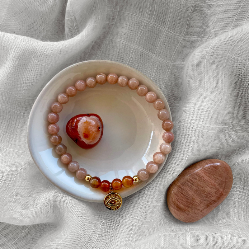 A bracelet made of peach moonstone beads, 4 carnelian beads and 2 small golden beads with a golden evil eye charm in the middle on a ceramic plate. In the middle of the plate is a carnelian gemstone. Next to the plate is a moonstone. All items are on a light fabric.