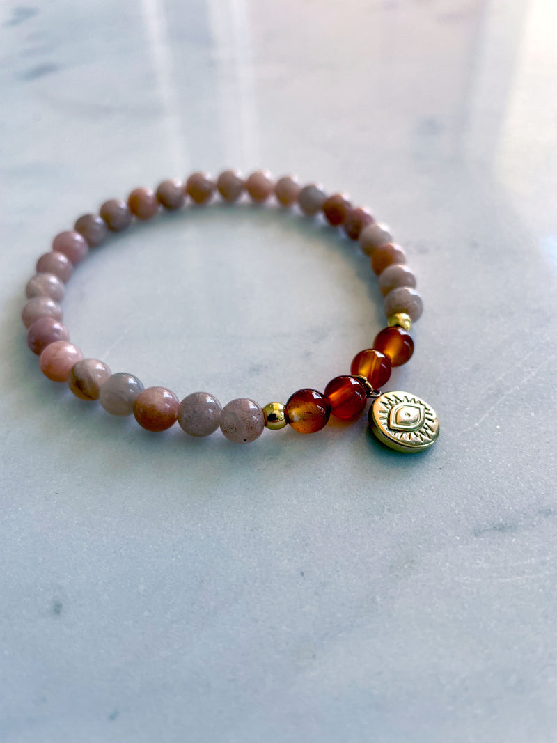 A bracelet made of peach moonstone beads, 4 carnelian beads and 2 small golden beads with a golden evil eye charm in the middle on a marble countertop.