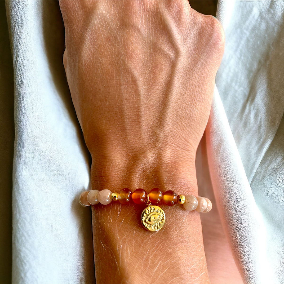 A bracelet made of peach moonstone beads, 4 carnelian beads and 2 small golden beads with a golden evil eye charm worn on a wrist.