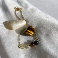 A close-up of a fine golden necklace with 2 citrine beads, 2 tiger eye beads and one golden bead in the middle on a light fabric. Next to the necklace is a tiger eye and a citrine gemstone.