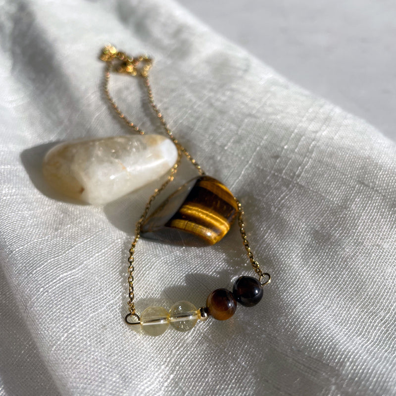 A close-up of a fine golden necklace with 2 citrine beads, 2 tiger eye beads and one golden bead in the middle on a light fabric. Next to the necklace is a tiger eye and a citrine gemstone.