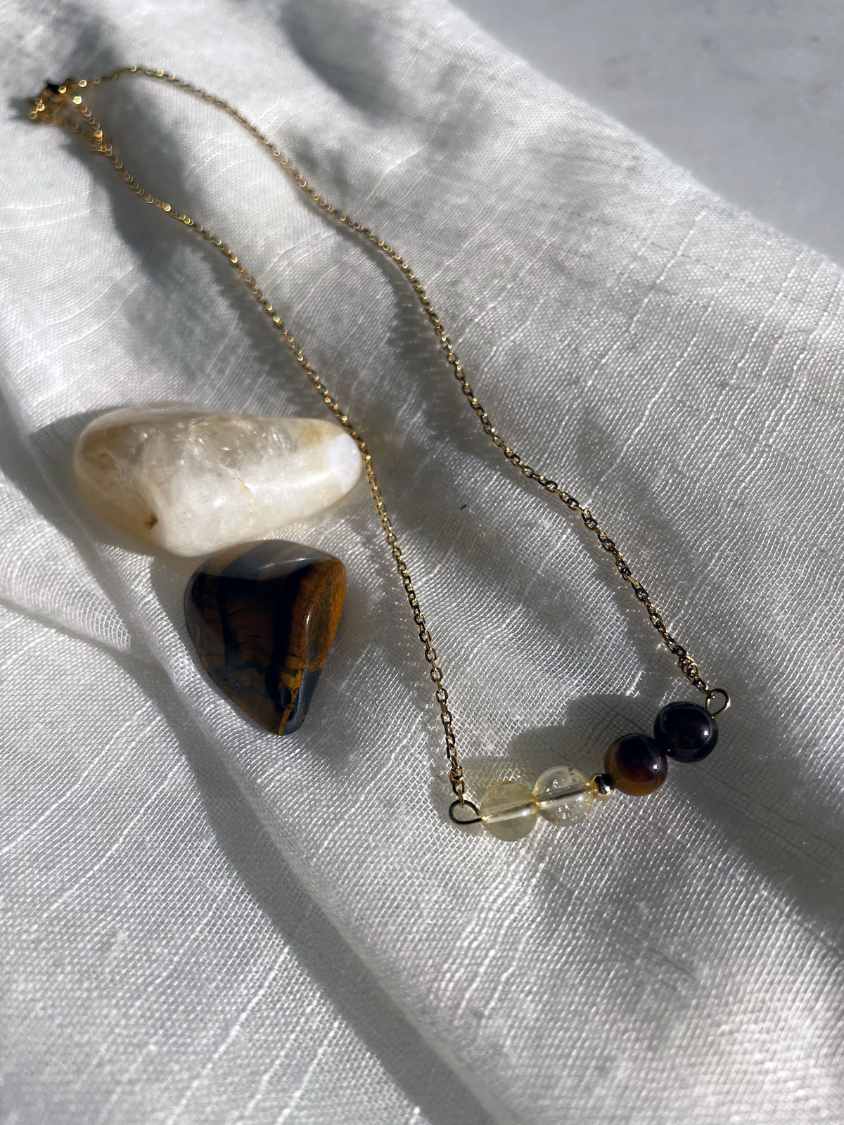 A fine golden necklace with 2 citrine beads, 2 tiger eye beads and one golden bead in the middle on a light fabric. Next to the necklace is a tiger eye and a citrine gemstone.