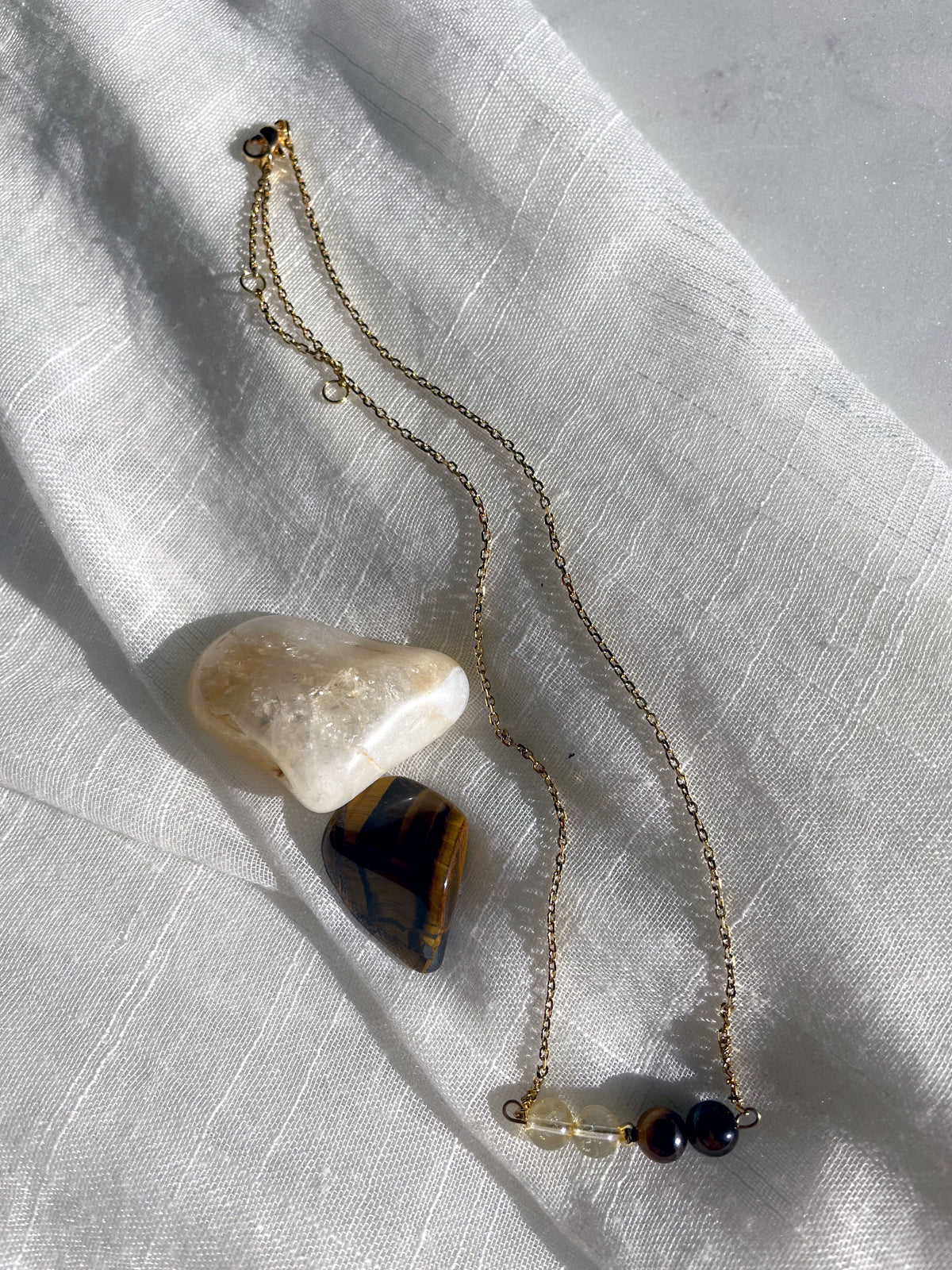 A fine golden necklace with 2 citrine beads, 2 tiger eye beads and one golden bead in the middle on a light fabric. Next to the necklace is a tiger eye and a citrine gemstone.