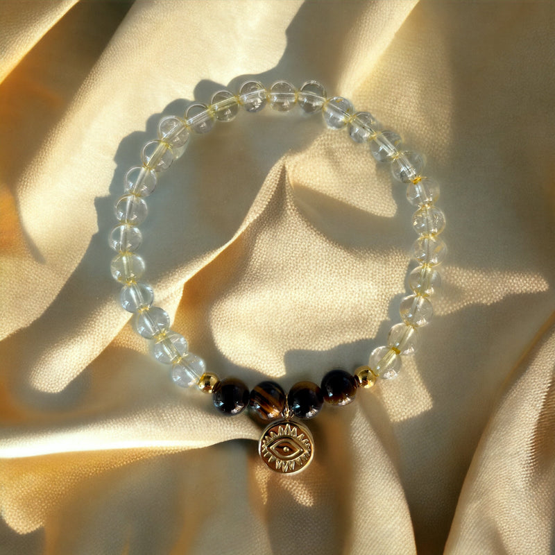 A bracelet made of citrine beads, 4 tiger eye beads and 2 small golden beads with a golden evil eye charm on a light yellow fabric.