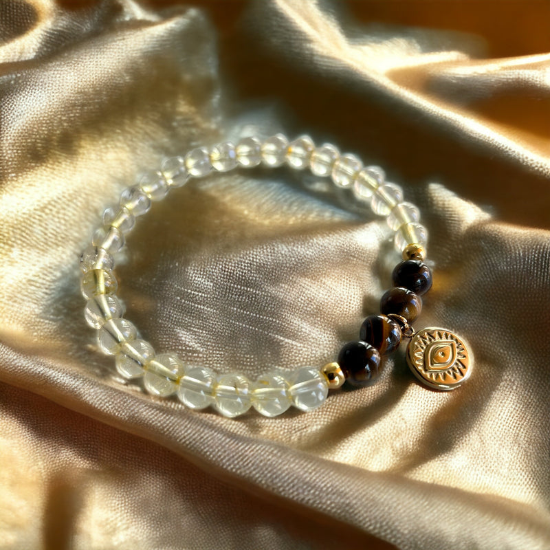 A bracelet made of citrine beads, 4 tiger eye beads and 2 small golden beads with a golden evil eye charm on a light yellow fabric.