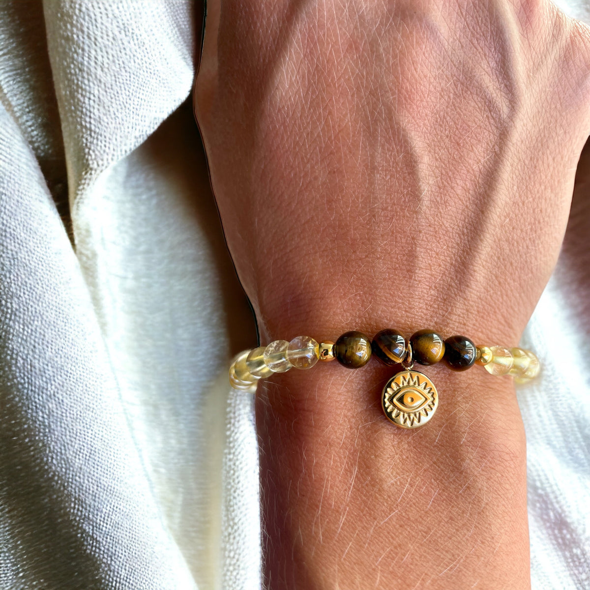 A bracelet made of citrine beads, 4 tiger eye beads and 2 small golden beads with a golden evil eye charm worn on a wrist.
