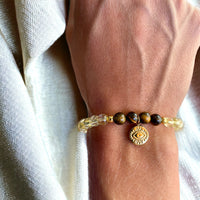 A bracelet made of citrine beads, 4 tiger eye beads and 2 small golden beads with a golden evil eye charm worn on a wrist.