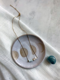 A fine golden necklace with 2 aquamarine beads, 2 white opal beads and one golden bead in the middle on a ceramic plate. Next to the plate is an Aquamarine gemstone.