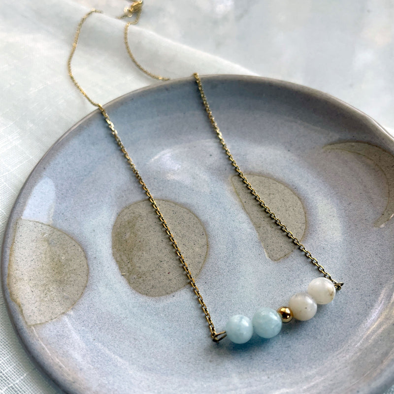A close-up of a fine golden necklace with 2 aquamarine beads, 2 white opal beads and one golden bead in the middle on a ceramic plate.