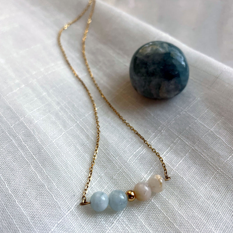 A close-up of a fine golden necklace with 2 aquamarine beads, 2 white opal beads and one golden bead in the middle on a on a light fabric next to an Aquamarine gemstone.