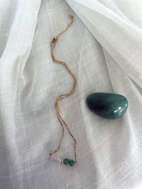 A fine golden necklace with 2 rose quartz beads, 2 green aventurine beads and one golden bead in the middle on a light fabric. Next to the necklace is a Green Aventurine gemstone.