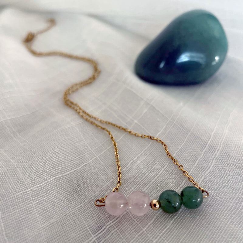 A close-up of a fine golden necklace with 2 rose quartz beads, 2 green aventurine beads and one golden bead in the middle on a light fabric. Next to the necklace is a Green Aventurine gemstone.