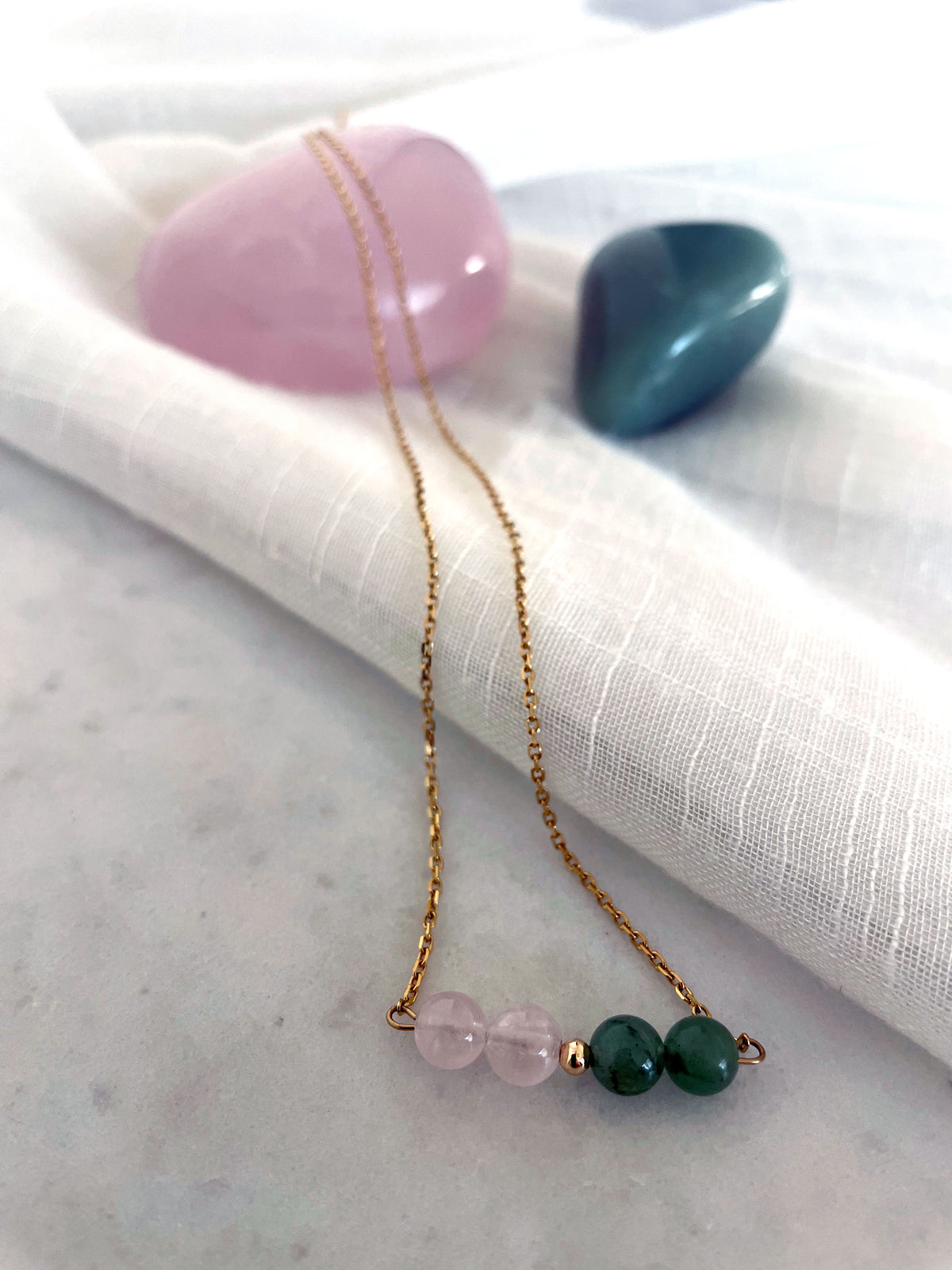 A fine golden necklace with 2 rose quartz beads, 2 green aventurine beads and one golden bead in the middle on a light fabric lying over a Rose Quartz gemstone. Next to the necklace is a Green Aventurine gemstone.