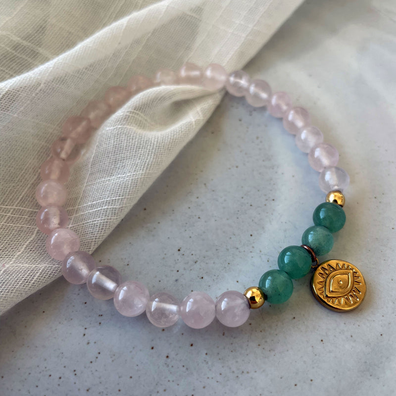 A bracelet made of rose quartz beads, 4 green aventurine beads and 2 small golden beads with a golden evil eye charm in the middle on a light fabric.