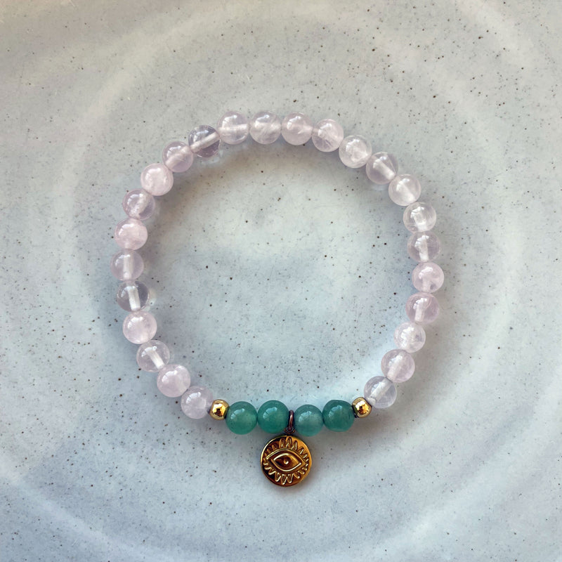 A bracelet made of rose quartz beads, 4 green aventurine beads and 2 small golden beads with a golden evil eye charm in the middle of a ceramic.