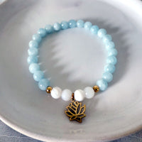 “I Speak My Truth” bracelet by Moksha Jewelry, featuring aquamarine and white opal beads with gold-plated accents and a lotus charm, displayed on a white ceramic dish—designed to support the throat chakra and encourage authentic communication and creativity.