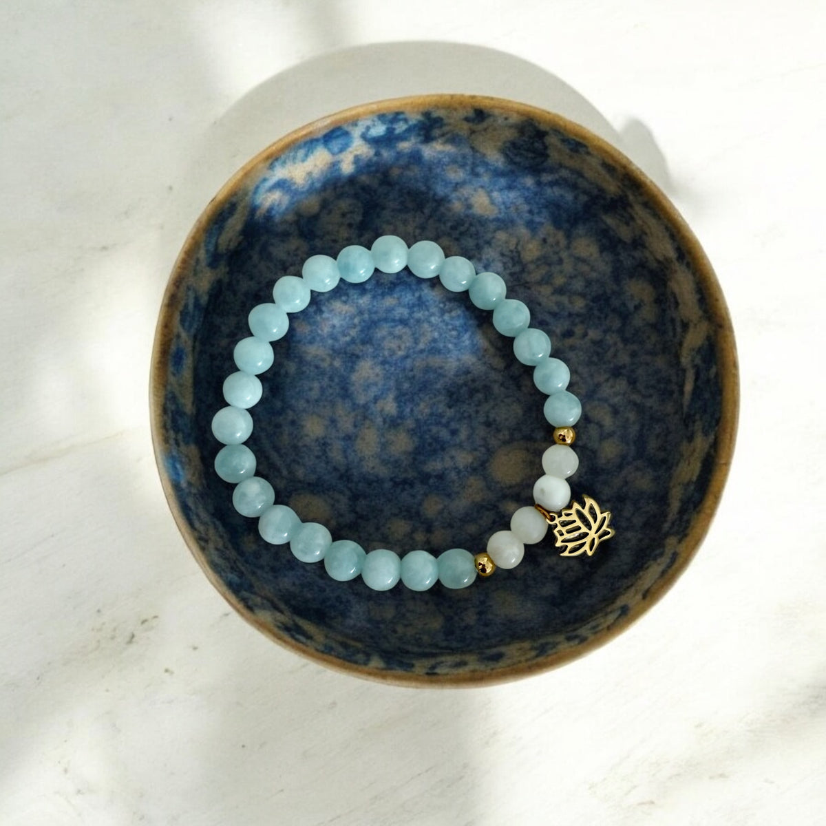 “I Speak My Truth” chakra bracelet by Moksha Jewelry, handcrafted with aquamarine and white opal beads, gold accents, and a lotus charm, displayed in a blue ceramic bowl—designed to activate the throat chakra and support clarity, creativity, and authentic expression.