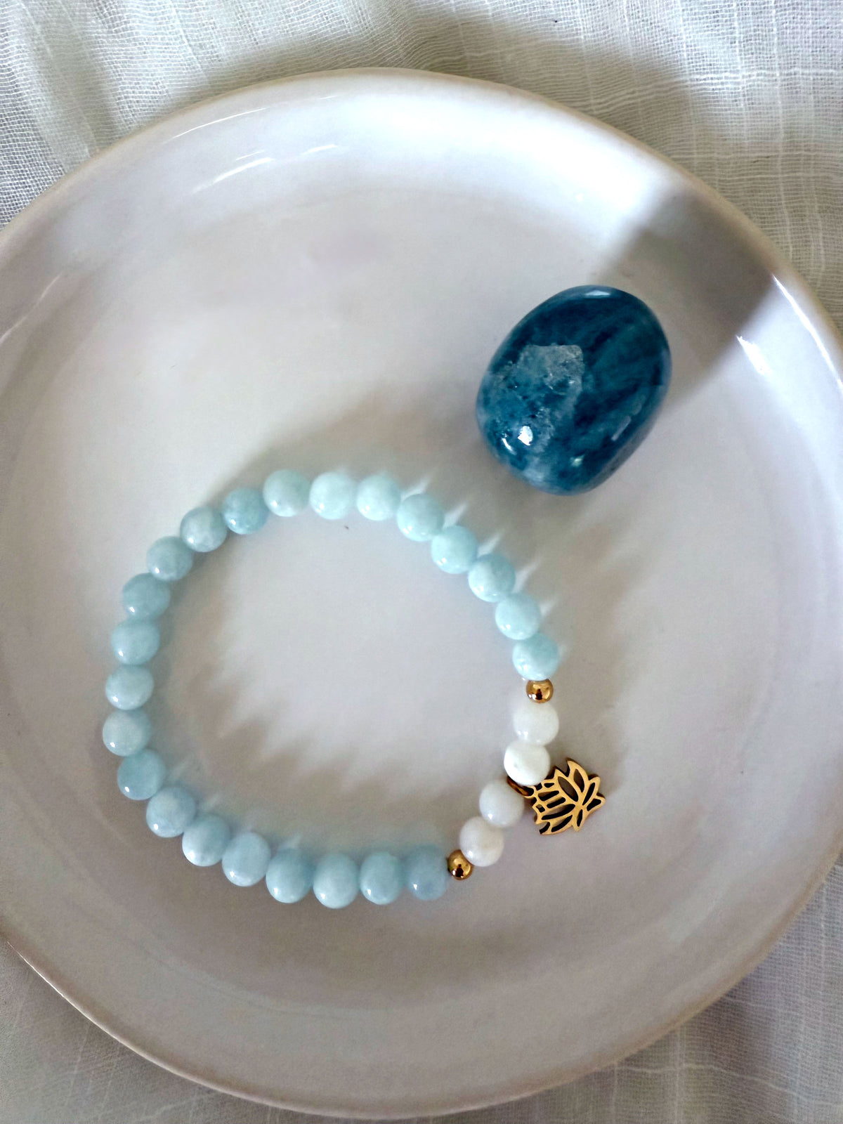 “I Speak My Truth” bracelet by Moksha Jewelry, handcrafted with aquamarine and white opal beads, accented with a gold lotus charm, displayed on a white ceramic dish beside a polished blue apatite stone—symbolizing throat chakra activation, clarity, and self-expression.