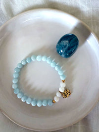 “I Speak My Truth” bracelet by Moksha Jewelry, handcrafted with aquamarine and white opal beads, accented with a gold lotus charm, displayed on a white ceramic dish beside a polished blue apatite stone—symbolizing throat chakra activation, clarity, and self-expression.