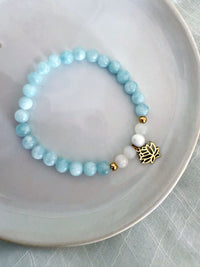 “I Speak My Truth” bracelet by Moksha Jewelry, featuring aquamarine and white opal gemstones with gold accents and a lotus charm, displayed on a smooth white ceramic plate—crafted to activate the throat chakra and support authentic, heartfelt expression.