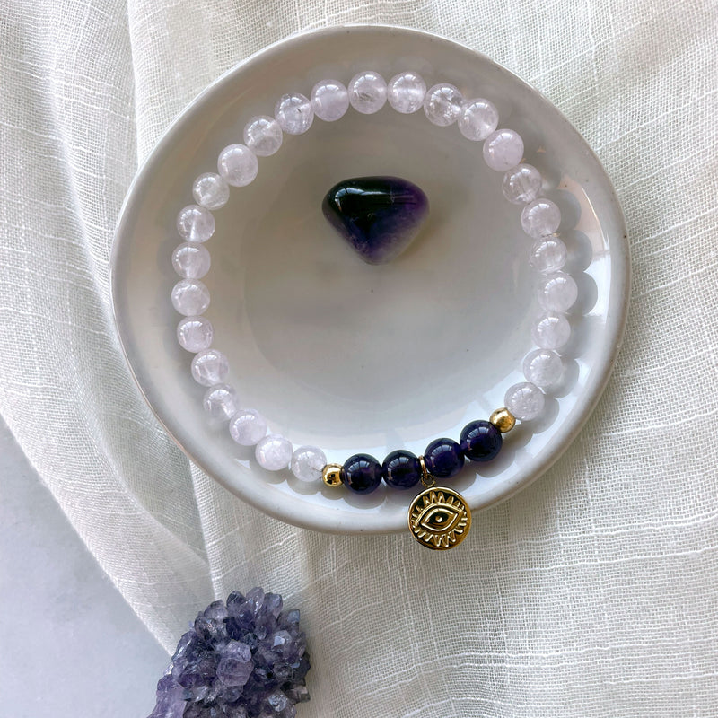 A bracelet made of lavender Amethyst and Amethyst beads, 2 small golden beads with a golden evil eye charm in the middle in a ceramic bowl on a light fabric with an Amethyst druse and a tumbled Amethyst.