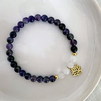 “Pure Consciousness” bracelet by Moksha Jewelry, made with deep amethyst and clear quartz beads, accented with gold-plated details and a lotus charm, displayed on a white ceramic plate—designed to activate the crown chakra and support spiritual clarity, balance, and awareness.