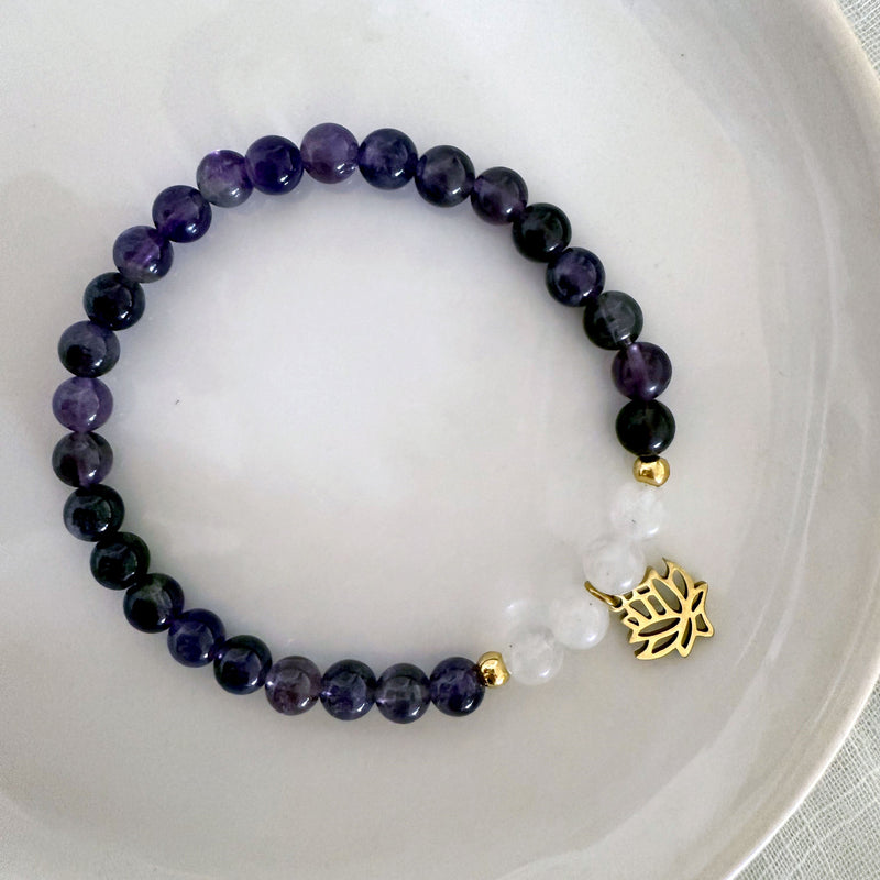 “Pure Consciousness” bracelet by Moksha Jewelry, made with deep amethyst and clear quartz beads, accented with gold-plated details and a lotus charm, displayed on a white ceramic plate—designed to activate the crown chakra and support spiritual clarity, balance, and awareness.